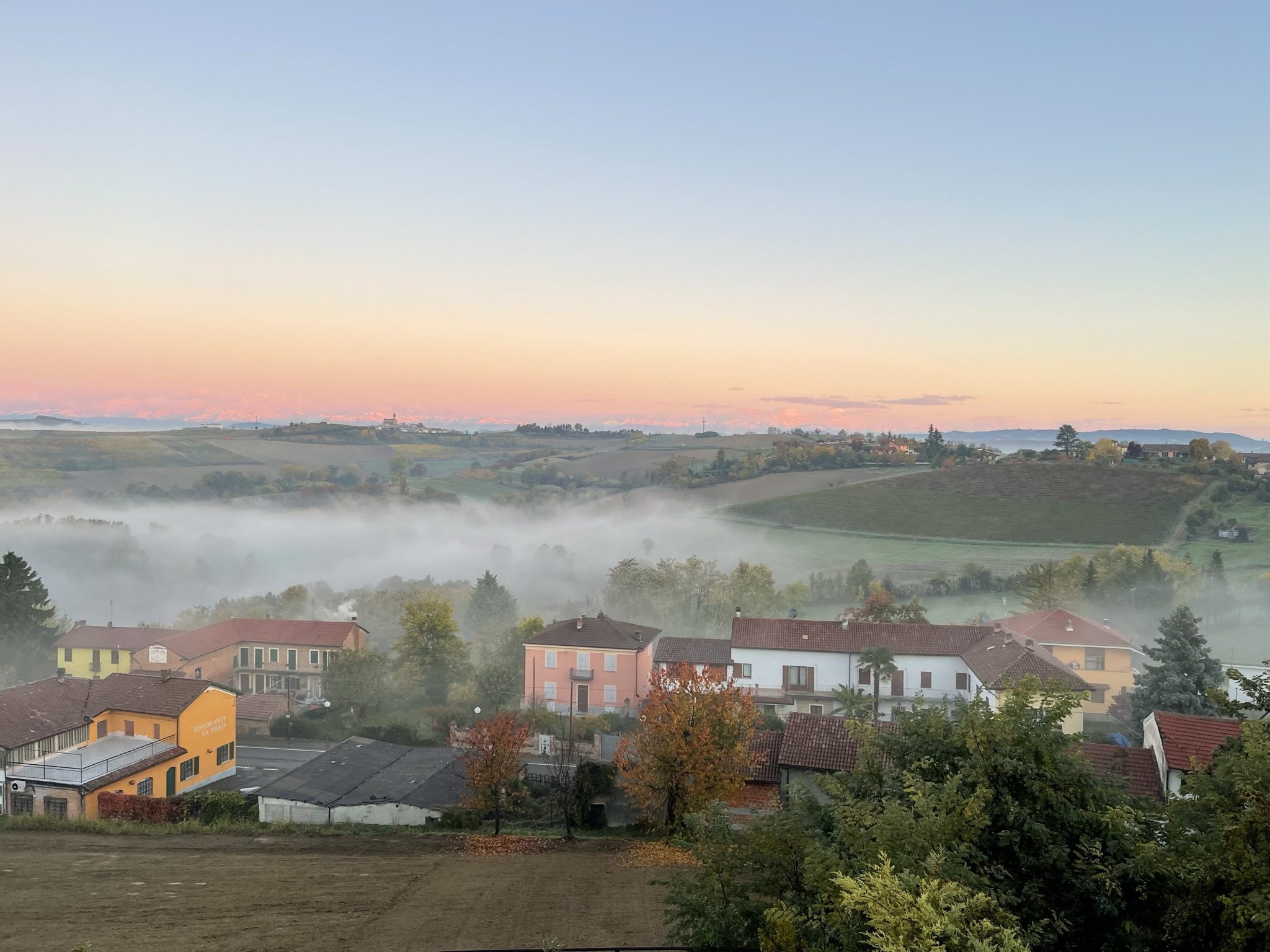 Dawn over Montemagno: mist filling the valley below terracotta rooftops, with the snow-covered Alps catching pink light on the horizon